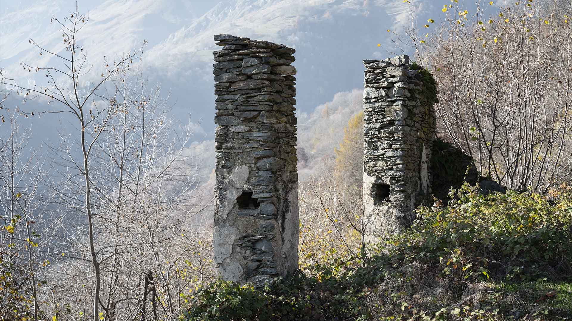 Ruderi di antichi pilastri in pietra a Ostana con bosco autunnale e cime alpine sullo sfondo